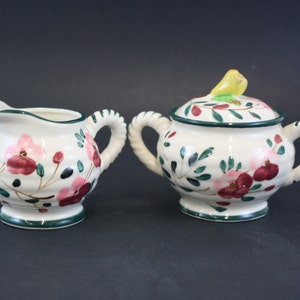 May include: A cream-colored ceramic creamer and sugar bowl set. The set features a floral design with pink and maroon flowers, green leaves, and black accents. The sugar bowl has a lid with a yellow bird-shaped handle. Both pieces have green trim and twisted handles.