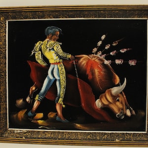 May include: A framed painting on black velvet depicting a matador in a yellow and blue suit, holding a red cape, facing a brown bull with a white horn. The bull is lying on the ground with several spears sticking out of its back.