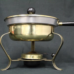 May include: A brass fondue pot with a black handle and a black base. The pot has a silver-colored lid with a black knob.