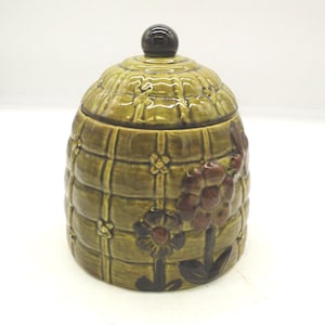 May include: A vintage ceramic jar with a lid. The jar is olive green with a textured, grid-like pattern and floral accents in brown and dark red. The lid has a dark brown knob.