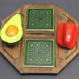 May include: An octagonal wooden serving board featuring two square green and black patterned tiles in the center. A three-dimensional painted avocado half is attached to the left side, and a red bell pepper is attached to the right side.