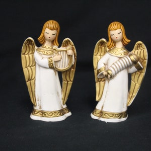 May include: Two white ceramic angel figurines with gold wings and accents. One angel is playing a harp and the other is playing a scroll.