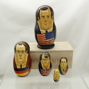 May include: A set of five wooden nesting dolls, each painted with a different portrait of a person. The largest doll is painted with a man wearing a suit and tie, holding an American flag. The other dolls are painted with men wearing suits and ties. The dolls are arranged in a row on a wooden surface.