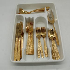 May include: A set of gold-colored flatware, including knives, forks, spoons, and serving utensils, arranged in a white plastic tray. The flatware has a textured, ornate design.