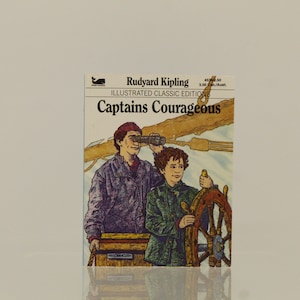 May include: A book cover with an illustration of two people on a boat. The book is titled "Captains Courageous" by Rudyard Kipling. The book is part of the Illustrated Classic Editions series.