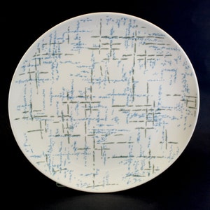 May include: A white ceramic dinner plate with a blue and green abstract pattern. The pattern is made up of thin lines and dots.