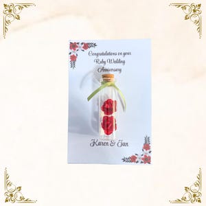 May include: A white card featuring a glass bottle with two red roses inside, tied with a green ribbon and cork stopper. Text reads "Congratulations on your Ruby Wedding Anniversary", signed "Karen & Jan". Floral accents.