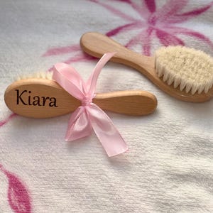 Puede incluir: Dos cepillos de pelo para bebé de madera con cerdas suaves. Un cepillo está grabado con el nombre "Kiara" y atado con una cinta rosa. Los cepillos están sobre una toalla blanca con un diseño floral rosa.