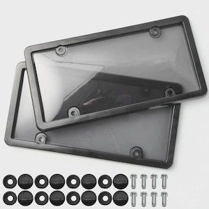 May include: Two black license plate frames with clear covers are stacked. The image also shows black screw caps, washers, and silver screws. The frames have rounded corners and mounting holes. The items are displayed on a light background.