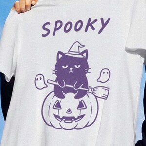 Puede incluir: Camiseta blanca con un gráfico morado de Halloween. El diseño presenta un gato negro con un sombrero de bruja, sentado en una calabaza, con la palabra "Spooky" arriba. También se incluyen dos fantasmas y una escoba.