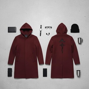 Puede incluir: Un abrigo con capucha color granate con un dise&ntilde;o bordado en negro en la espalda, exhibido con una cartera negra, tel&eacute;fono, reloj, gafas, auriculares, gorro y una taza. El abrigo es de estilo largo y entallado.