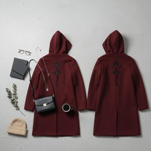 Puede incluir: Dos abrigos con capucha burdeos con dise&ntilde;os bordados en negro en la espalda. Un bolso negro, un collar dorado, un gorro beige y una taza de caf&eacute; negra est&aacute;n colocados alrededor de un abrigo. Los abrigos son de estilo largo y entallado.