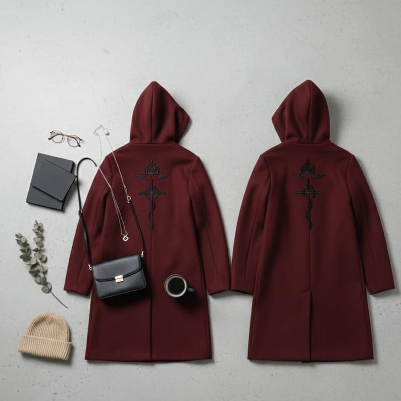 Puede incluir: Dos abrigos con capucha burdeos con dise&ntilde;os bordados en negro en la espalda. Un bolso negro, un collar dorado, un gorro beige y una taza de caf&eacute; negra est&aacute;n colocados alrededor de un abrigo. Los abrigos son de estilo largo y entallado.