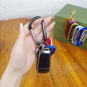 May include: A black metal carabiner keychain engraved with the text Merry Christmas! attached to a black car key fob. Additional colorful carabiners in red, green, purple, yellow, and blue are arranged in a row on a green box in the background.