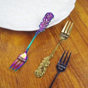 May include: Three decorative forks with floral designs. One fork has a rainbow finish, another is gold, and the third is black. Each fork has the words "Custom Text" engraved on it. The forks are placed on a wooden surface.