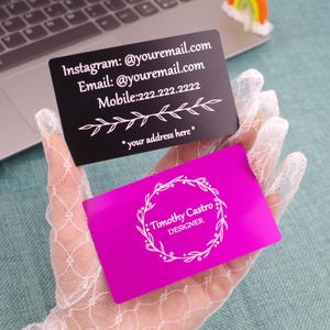 Puede incluir: Dos tarjetas de visita, una negra y otra magenta, se sostienen en una mano. La tarjeta negra tiene texto blanco, incluyendo nombres de redes sociales y un número de teléfono. La tarjeta magenta presenta el nombre y el título de un diseñador.