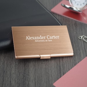 Może przedstawiać: Etui na wizytówki w kolorze różowego złota z napisem "Alexander Carter" i tytułem "Attorney at law" wydrukowanym na biało. Etui ma szczotkowane metalowe wykończenie i zawiasowe zamknięcie.