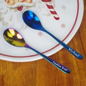 Puede incluir: Dos cucharas iridiscentes con acabado azul y dorado descansan sobre un plato con temática navideña. Una cuchara está grabada con "Michael" y la otra con "Custom Text". Las cucharas están sobre una superficie de madera.