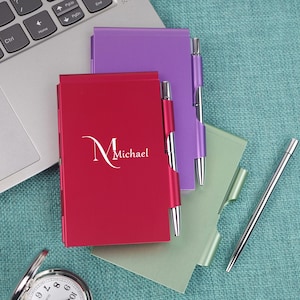May include: A red, purple, and green notepad with a pen holder and a silver pen. The red notepad has the name "Michael" printed on it. The notepads are on a teal surface next to a laptop.