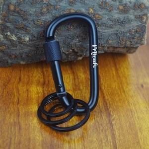 May include: Black carabiner with a key ring, featuring the name "Micah" in white script. The carabiner is made of metal and has a textured locking mechanism. The key ring holds several small black rings.