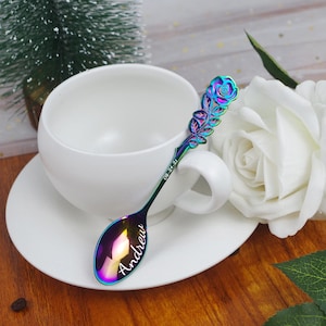 May include: A rainbow-colored metal spoon with a rose-shaped handle, featuring the name Andrew engraved on the bowl and the date 08-24-21 engraved on the stem, resting against a white ceramic teacup and saucer set with a white rose nearby.
