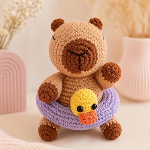 May include: A handmade crochet bear in shades of brown and tan, with a brown snout and paws. It's wearing a purple floatie with a yellow duck attached. A charming, whimsical toy, perfect for a child's room.