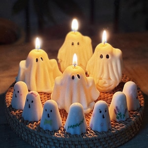May include: A collection of ghost-shaped candles arranged on a round woven tray. Four larger candles are lit with visible flames, while several smaller, unlit ghost figurines with painted details like bows and flowers surround them.