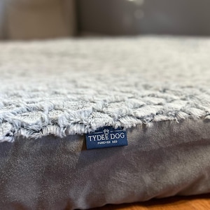 May include: A rectangular pet bed featuring a textured, light gray faux fur top surface and smooth, dark gray sides. A small blue fabric tag is attached to the side, displaying the white text TYDEE DOG FUREVER BED with a small paw print icon.