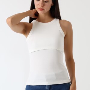 K&ouml;nnte beinhalten: Ein wei&szlig;es geripptes Tanktop mit einem geschichteten Design. Das Oberteil hat einen runden Ausschnitt und eine figurbetonte Silhouette. Das Model tr&auml;gt eine blaue Jeans. Der Gesamtstil ist l&auml;ssig und modern.