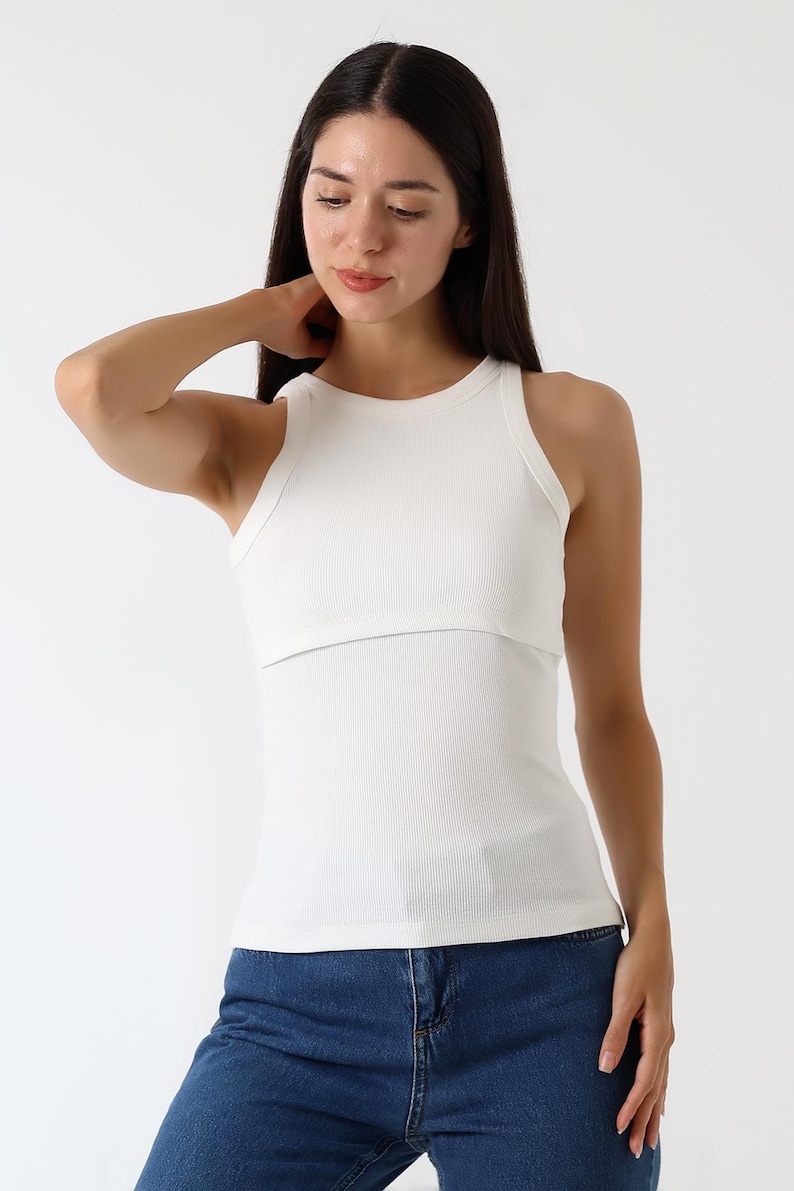 K&ouml;nnte beinhalten: Ein wei&szlig;es geripptes Tanktop mit einem geschichteten Design. Das Oberteil hat einen runden Ausschnitt und eine figurbetonte Silhouette. Das Model tr&auml;gt eine blaue Jeans. Der Gesamtstil ist l&auml;ssig und modern.