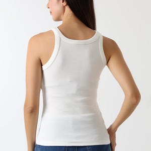 K&ouml;nnte beinhalten: Ein wei&szlig;es geripptes Tanktop mit Rundhalsausschnitt und breiten Tr&auml;gern. Das Tanktop wird mit blauen Jeans getragen. Das Model ist von hinten zu sehen.