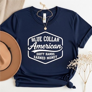 May include: A navy blue t-shirt featuring the white text "BLUE COLLAR AMERICAN" and "DIRTY HANDS EARNED MONEY". A brown hat and gold necklace are also visible in the image. The t-shirt is knotted at the bottom.