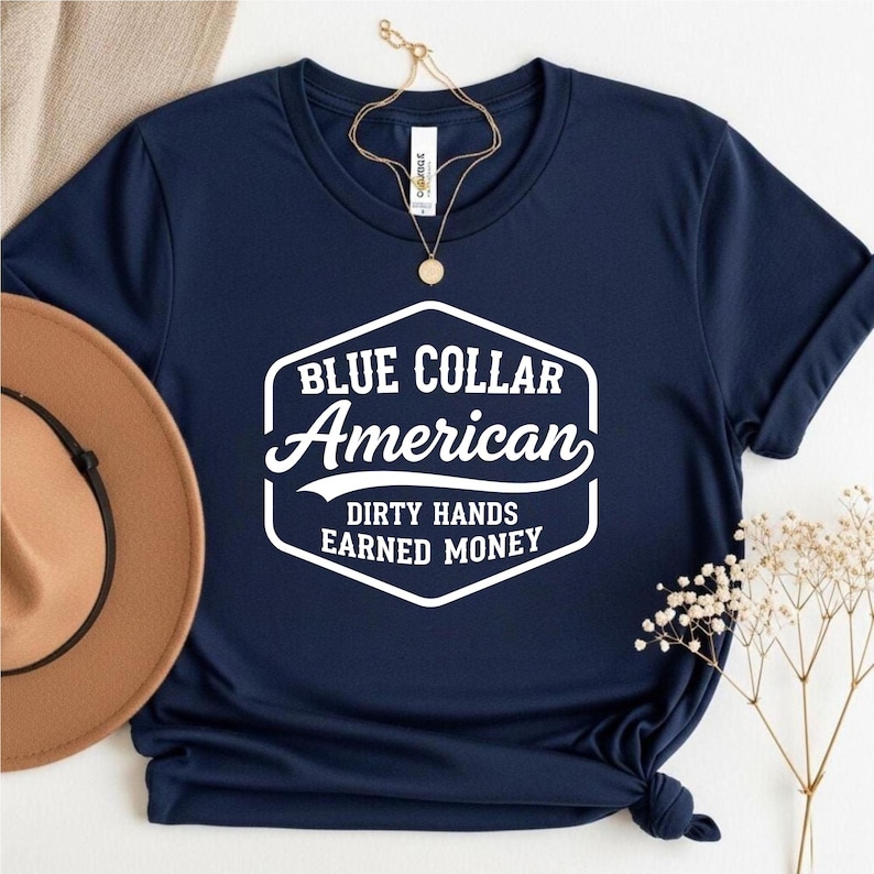 May include: A navy blue t-shirt featuring the white text "BLUE COLLAR AMERICAN" and "DIRTY HANDS EARNED MONEY". A brown hat and gold necklace are also visible in the image. The t-shirt is knotted at the bottom.