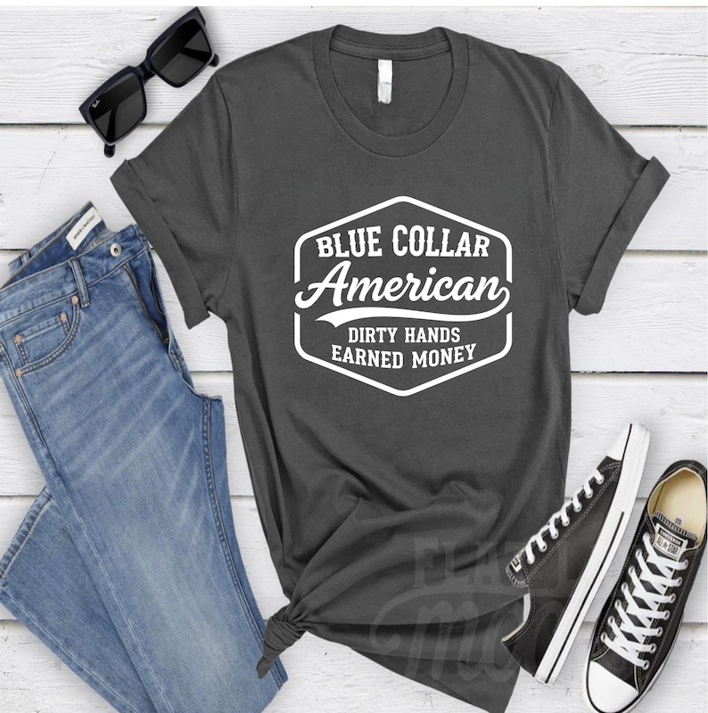 May include: Dark grey t-shirt with white text reading "BLUE COLLAR American DIRTY HANDS EARNED MONEY". The shirt is styled with blue jeans, black sunglasses, and black trainers.