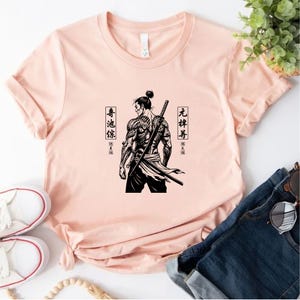 May include: A light pink t-shirt featuring a black and white illustration of a samurai with a sword on his back. The samurai has tattoos and is facing away from the viewer. Japanese characters are on either side of the samurai.