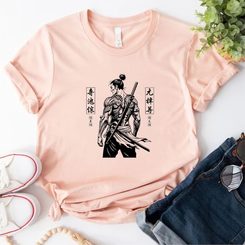 May include: A light pink t-shirt featuring a black and white illustration of a samurai with a sword on his back. The samurai has tattoos and is facing away from the viewer. Japanese characters are on either side of the samurai.