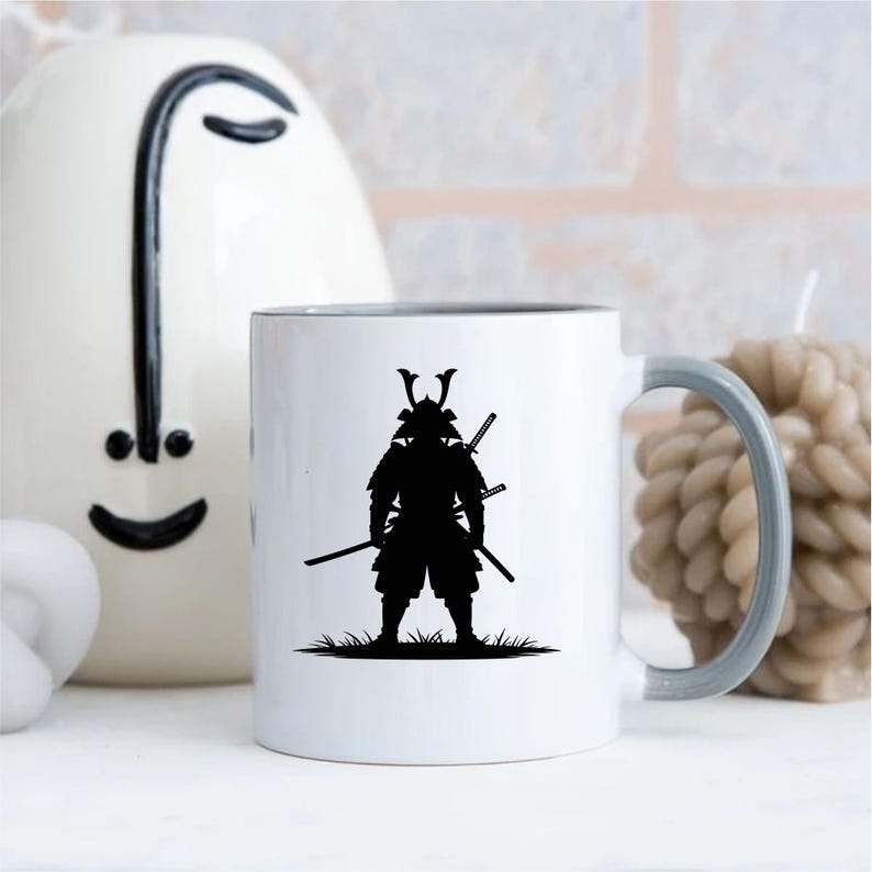 May include: White ceramic mug with a gray handle and rim, featuring a black silhouette of a samurai warrior with two swords. The design is set against a white background, with a decorative vase and candle in the background.