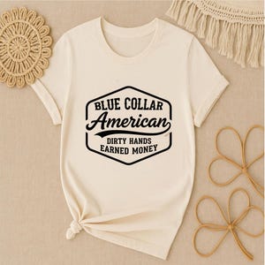 May include: A cream-coloured t-shirt with a black graphic that reads "BLUE COLLAR American" and "DIRTY HANDS EARNED MONEY". The shirt has short sleeves and a crew neck. The shirt is knotted at the bottom.