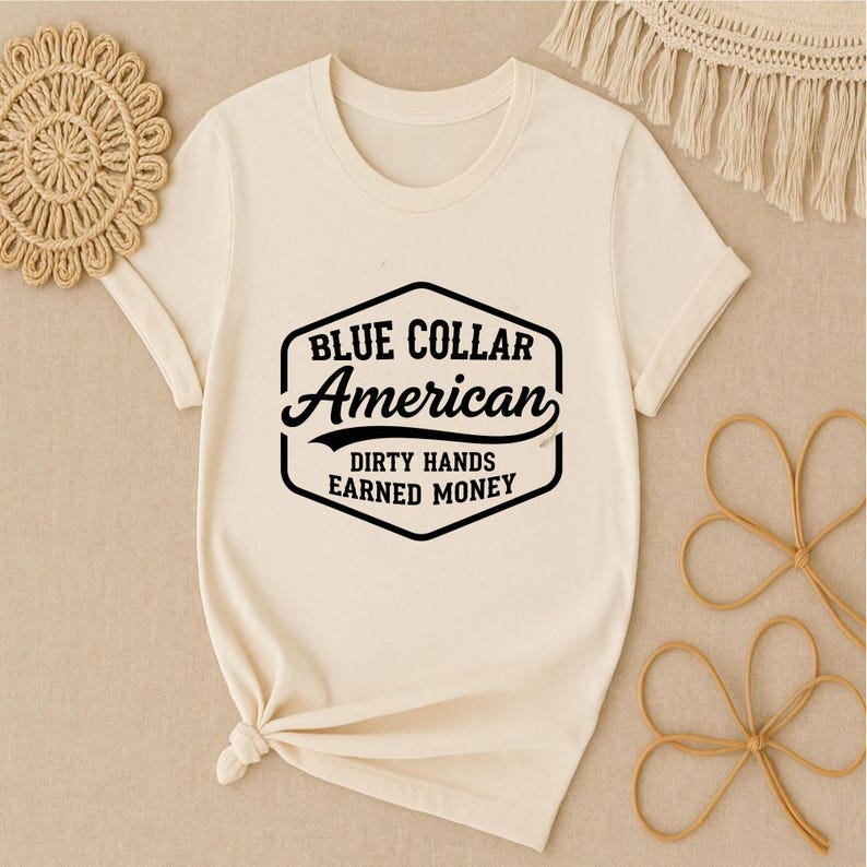 May include: A cream-coloured t-shirt with a black graphic that reads "BLUE COLLAR American" and "DIRTY HANDS EARNED MONEY". The shirt has short sleeves and a crew neck. The shirt is knotted at the bottom.