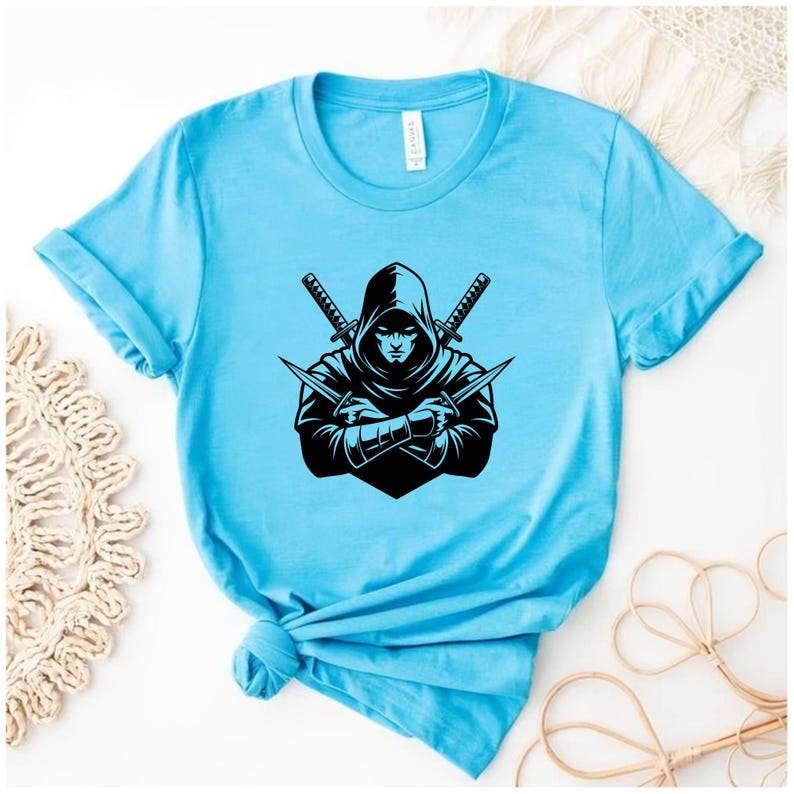 May include: Light blue t-shirt featuring a black graphic of a hooded figure with crossed swords and knives. The design is centered on the shirt, with the sleeves rolled up. The shirt is made of a soft material.