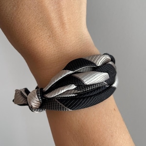 Può includere: Bracciale intrecciato con una combinazione di cordoncini neri e grigio argento. Il bracciale presenta un motivo spesso e strutturato, creando un accessorio audace ed elegante. Il design è moderno e adatto per un polso di circa 17 cm.