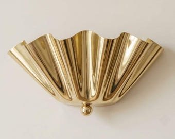 Könnte beinhalten: Eine Wandleuchte aus poliertem, goldfarbenem Metall mit einem breiten, halbkreisförmigen, geriffelten muschelartigen Design mit tiefen vertikalen Rillen. Die Leuchte ist in der unteren Mitte mit einer kleinen, glatten, kugelförmigen Akzentkugel versehen.