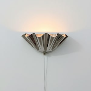 May include: A polished silver wall sconce with a scalloped, fan-like design. The light fixture is mounted on a white wall and has a small, round base. A thin, white cord hangs below the sconce.