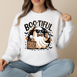 Puede incluir: Sudadera blanca con un diseño de Halloween. El diseño presenta un fantasma de dibujos animados con gafas de sol y pajarita, sosteniendo una bebida y una bolsa con estampado de leopardo. El texto "BOO-TIFUL" está encima del fantasma.