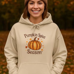 May include: A beige hoodie with a drawstring and a front pocket. The hoodie features the text "Pumpkin Spice Season" with a graphic of an orange pumpkin and autumn leaves. A brown beanie is worn.