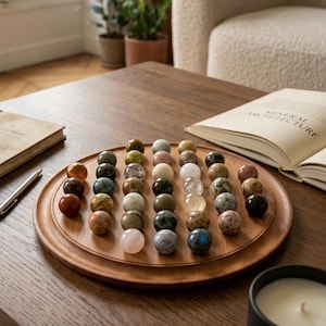 Könnte beinhalten: Ein Holztablett zeigt eine Sammlung polierter Edelsteine in verschiedenen Farben, darunter Rot, Grün und Weiß. Das Tablett steht auf einem dunklen Holztisch, mit einem Buch und einem Stift in der Nähe. Eine schwarze Kerze und ein beiger Sessel sind ebenfalls zu sehen.