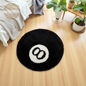 May include: A circular black rug featuring a white number eight design. The rug is made of a soft, plush material and is placed on a light-coloured wooden floor. The rug is a decorative item for a games room or a fun addition to a home.
