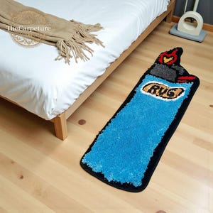 Peut inclure: Un tapis fantaisie en forme de briquet bleu avec le mot "RUG" dans un ovale jaune. Le tapis a une bordure noire et une surface texturée et hirsute. Le haut du briquet présente des détails gris, rouges et jaunes.