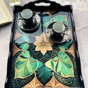 May include: A black serving tray with a floral design in shades of green, teal, and gold. Two black teacups and saucers with silver accents sit on the tray. The tray has handles and a glossy finish.