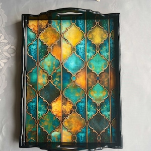 May include: A rectangular decorative tray with a black frame and handles. The tray's surface features a colorful geometric pattern in shades of teal, gold, and orange, with a dark outline. The tray is designed for serving or display.